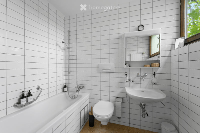 Modern bathroom with white tiled walls and floor, featuring a bathtub, toilet, sink, mirror, and showerhead.