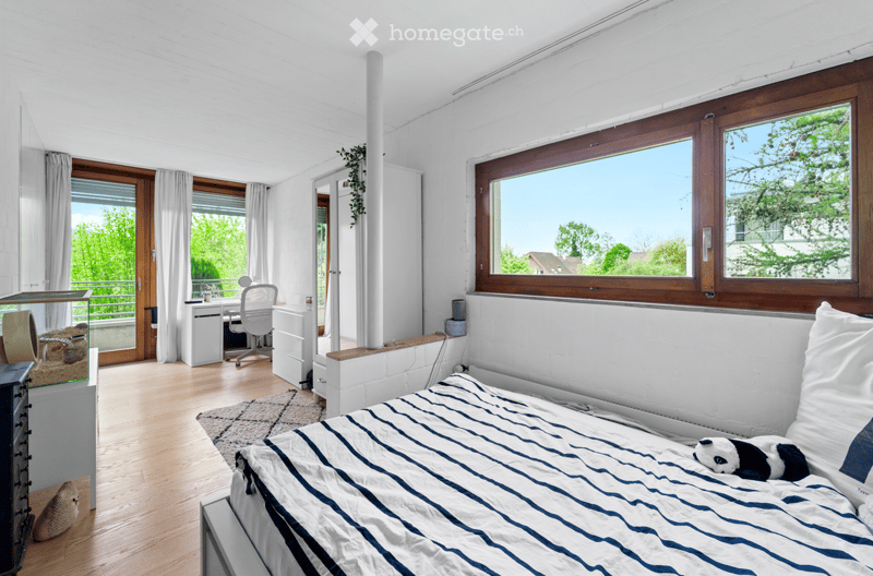 Bright bedroom with large windows, a bed with blue and white striped bedding, a desk with a chair, a small cabinet, and a balcony access.