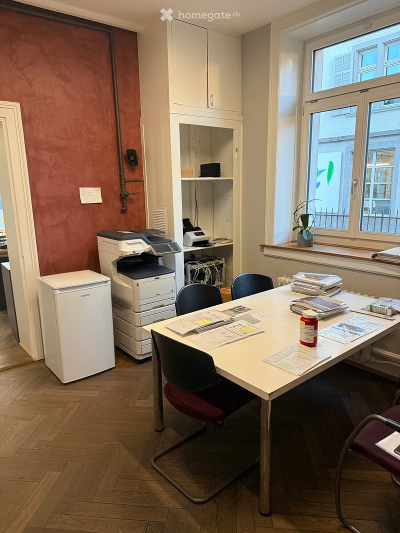 Office room with wooden floor, red wall, a white desk, printer on the floor, a small potted plant on the window sill, cabinets, chairs, table, papers, books and a red bottle