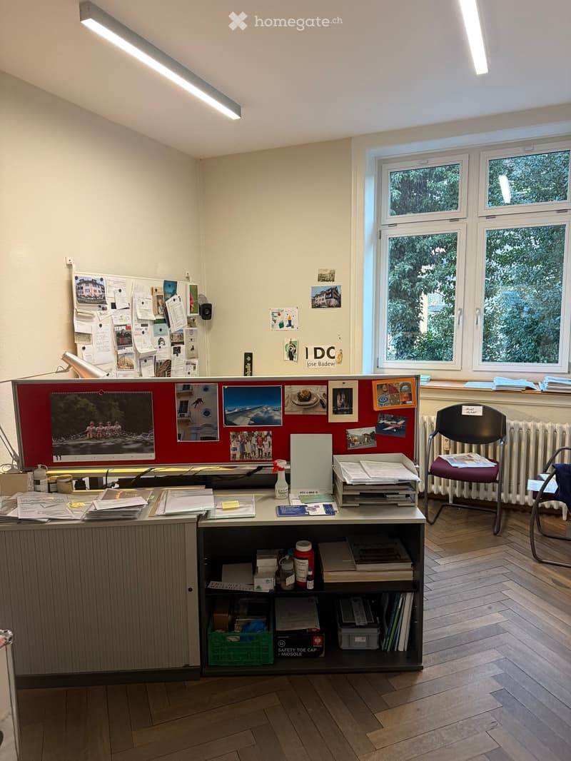 large desk with various items, bulletin board with papers and pictures, radiator, wooden floor, window with trees outside