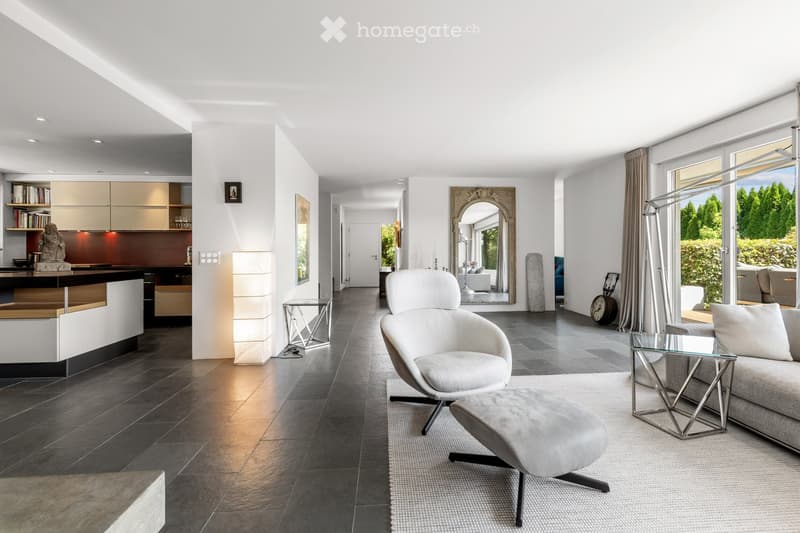 Modern living room with a grey sofa, white armchair, ottoman, coffee table, and a sleek kitchen area with a bar counter and bookshelf. Large windows and a mirror add brightness and depth to the space.