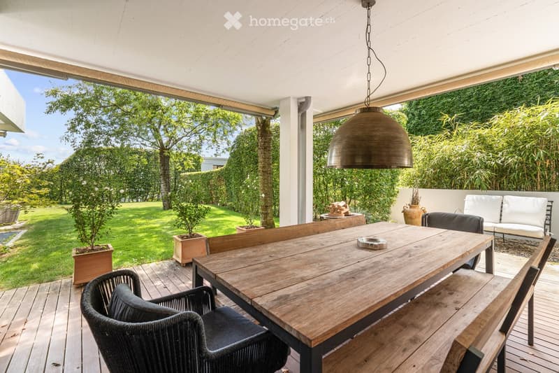 A covered outdoor terrace with a wooden dining table, wicker chairs, and a white sofa, surrounded by greenery and a well-maintained lawn.