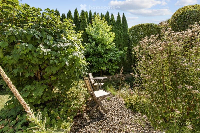 A lush garden with tall trees, flowering plants, and a small seating area featuring a wooden chair and table.