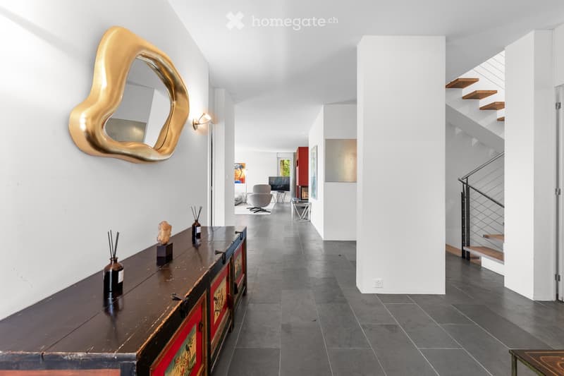 Modern hallway with white walls, dark tiled floor, a large gold-framed mirror, a wooden console table with decorative items, and a staircase leading upwards.