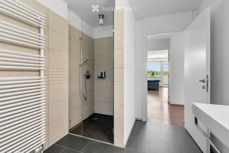 Modern bathroom with a glass-enclosed shower, a white radiator, a sink, and a view into an adjacent room with a bed and large windows.