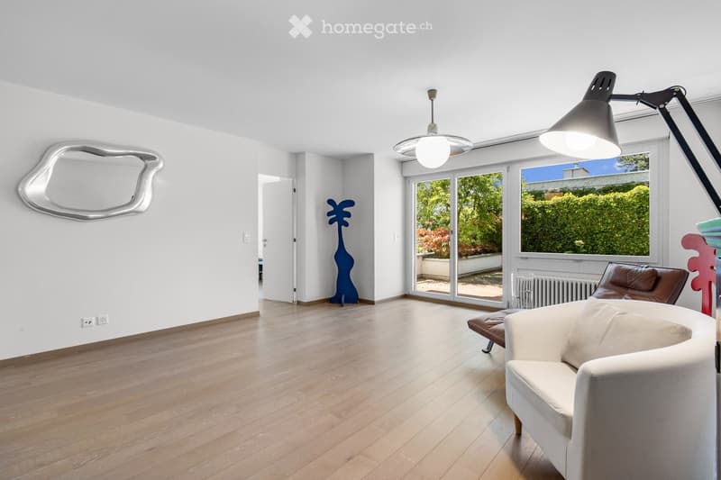 Bright living room with wooden flooring, large windows offering a view of greenery, a white sofa, a brown leather chair, a floor lamp, a table lamp, a decorative mirror, and a blue abstract sculpture.