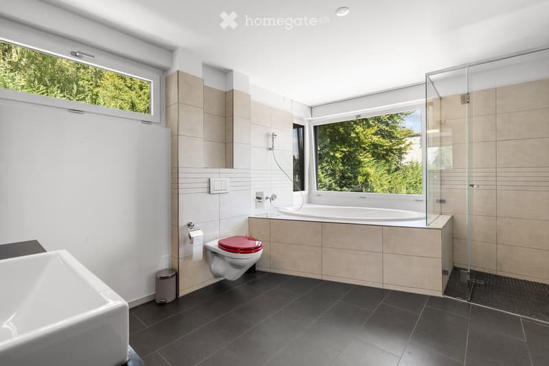 Modern bathroom with a large window, bathtub, toilet, and glass-enclosed shower. Features dark tiled flooring and light-colored walls.