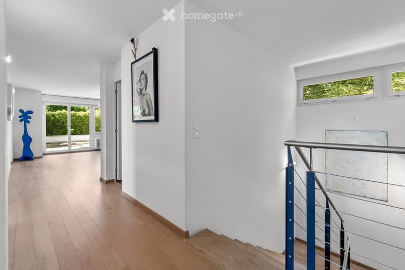 Bright hallway with wooden flooring, white walls, and large windows. Features a modern staircase with a metal railing and a framed artwork on the wall.