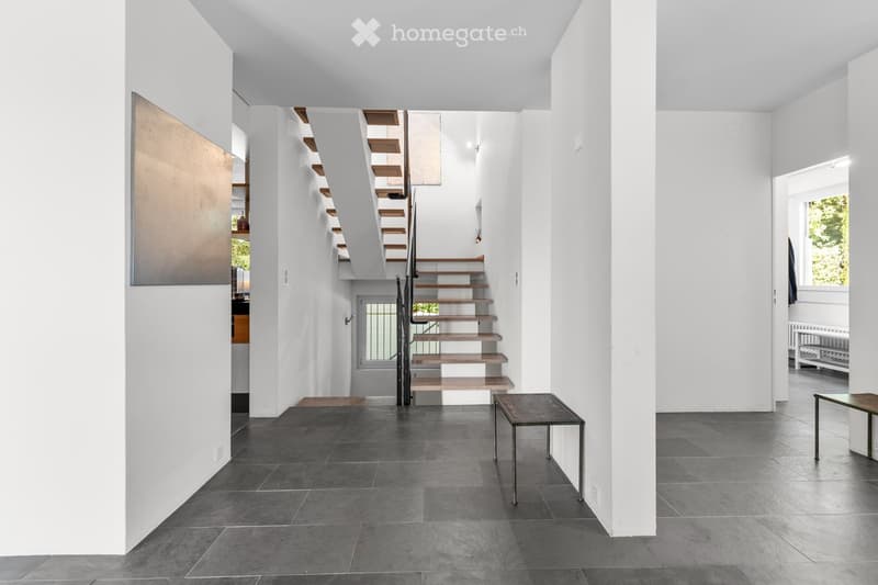 Modern, minimalist hallway with a sleek staircase featuring wooden steps and a metal railing, grey tiled floor, white walls, and a small metal side table.