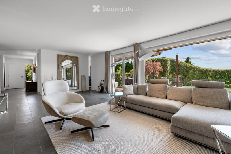 Modern living room with large windows offering a view of the garden, featuring a sectional sofa, armchair, ottoman, coffee table, and a floor lamp.