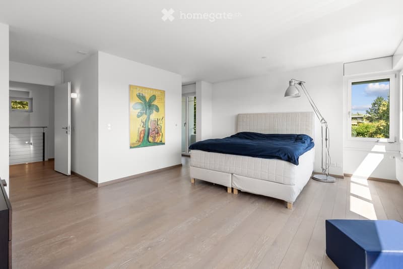 Bright, modern bedroom with wooden flooring, white walls, a large bed with a dark blue duvet, a floor lamp, and a colorful artwork on the wall.