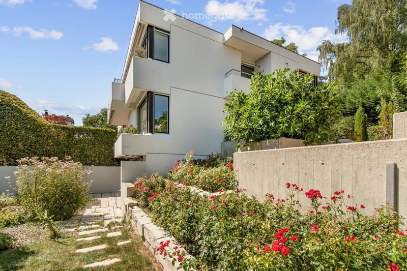 Modern two-story house with white exterior, large windows, and a raised ground floor. The garden features red flowers, a concrete wall, and a stone pathway.