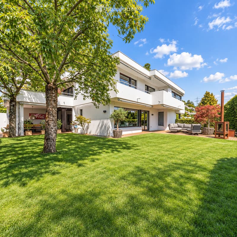 Modern white house with large windows and a balcony, surrounded by a well-maintained lawn, trees, and outdoor seating area.