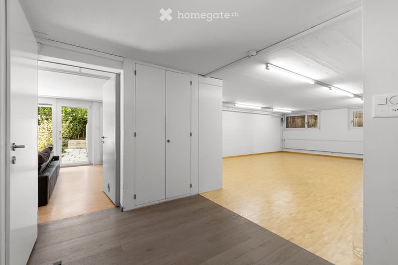 Modern, minimalist room with light wooden flooring, white walls, and a large open space. A black leather sofa is visible in the foreground, and there is a sliding glass door leading to an outdoor area with greenery.