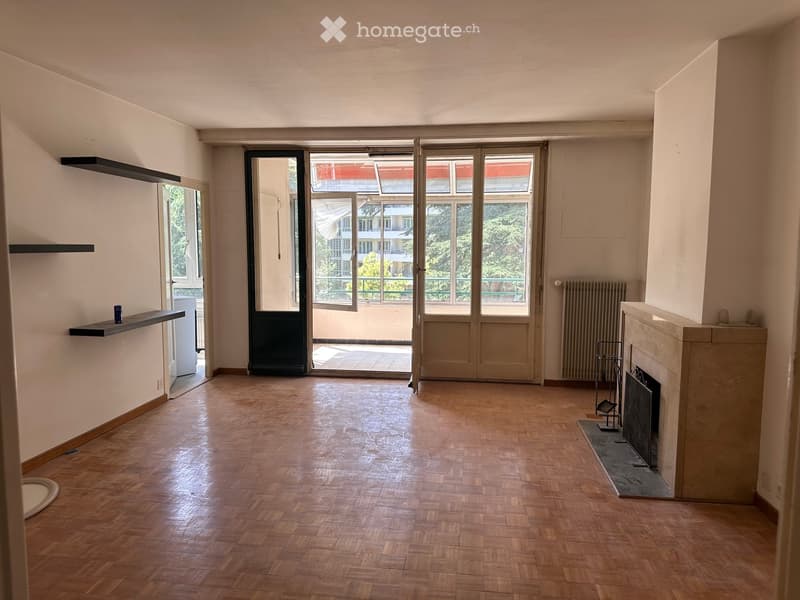 Empty room with wooden flooring, white walls, fireplace, large windows, and a balcony door leading outside.