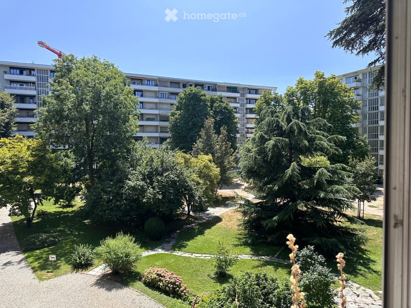 A well-maintained garden area with green lawns, trees, shrubs, and a gravel pathway, surrounded by apartment buildings.