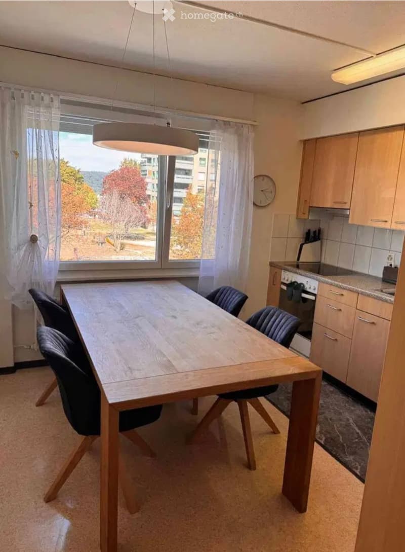 A modern dining area with a wooden dining table, four chairs, and a kitchen area with cabinets and an oven. The room features large windows with sheer curtains and a view of trees and buildings outside.