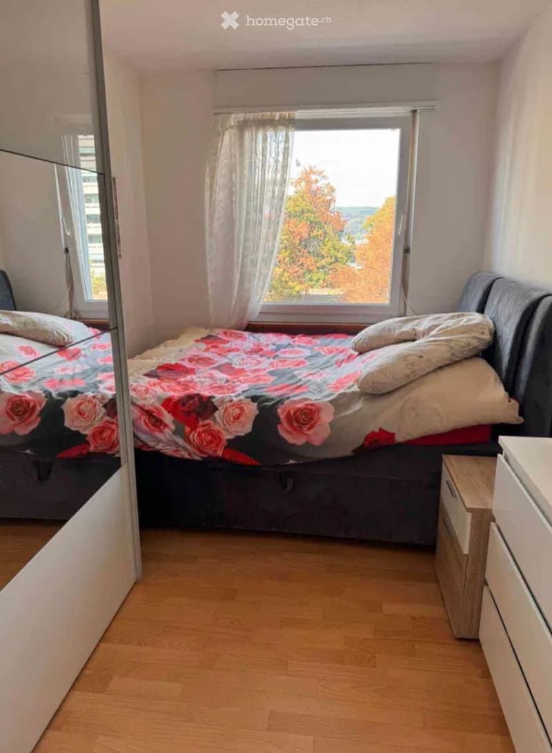 Cozy bedroom with wooden flooring, a large window with sheer curtains, a double bed with a floral duvet, and a nightstand with drawers.