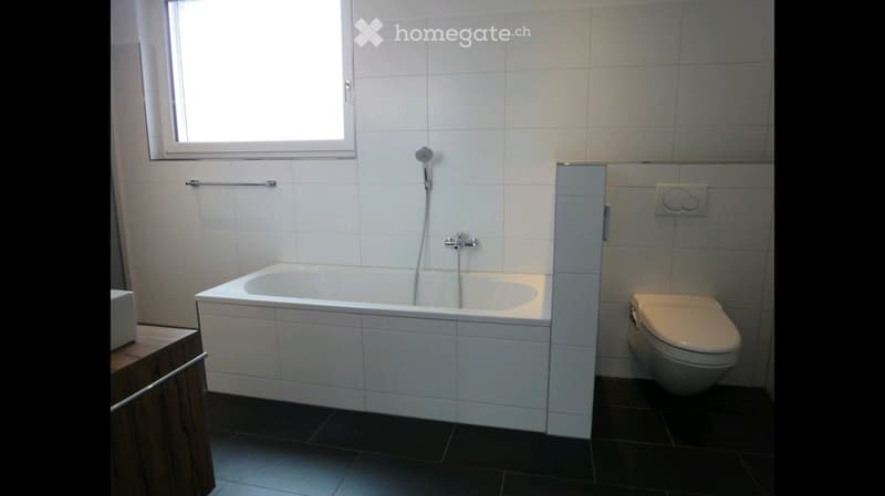 Modern bathroom with a white bathtub, toilet, and showerhead. The walls are tiled, and there is a towel rack on the wall.