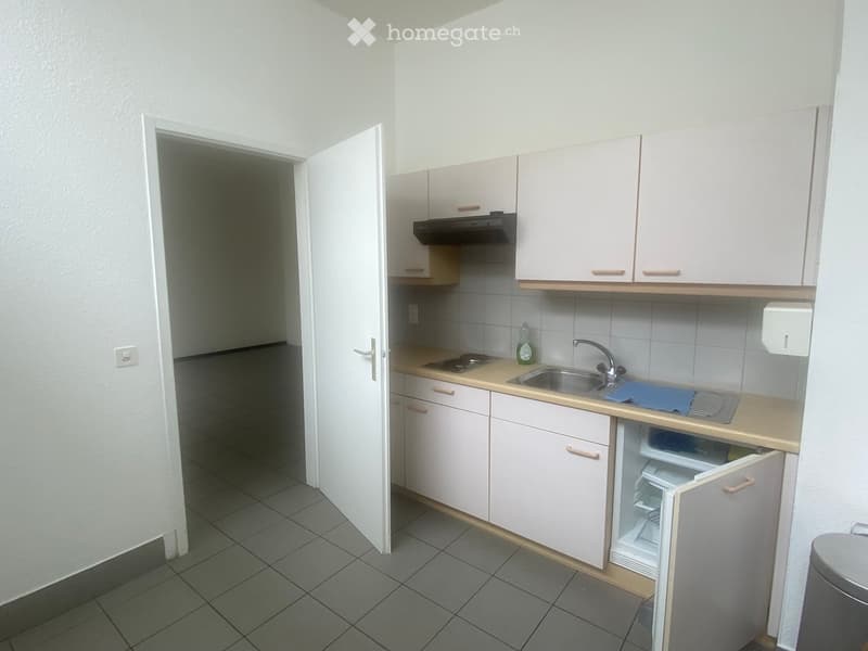 Compact kitchen with light-colored cabinets, a sink, a small refrigerator, and a stove. The space is clean and organized with tiled flooring and a doorway leading to another room.