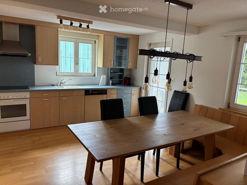 Bright kitchen with wooden cabinets, a large wooden dining table, four black chairs, and modern appliances including an oven and a sink.