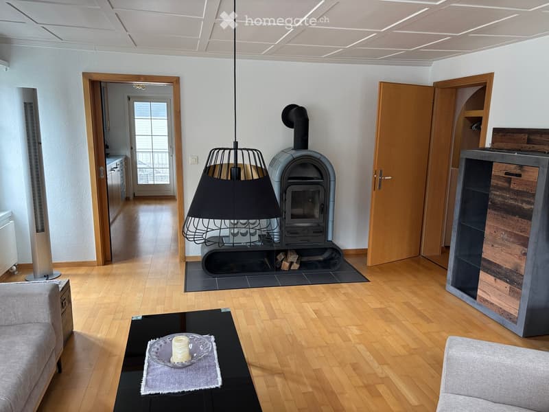 Modern living room with wooden flooring, a central pendant lamp, a grey sofa, a black coffee table with a decorative item, and a wood-burning stove with a chimney.