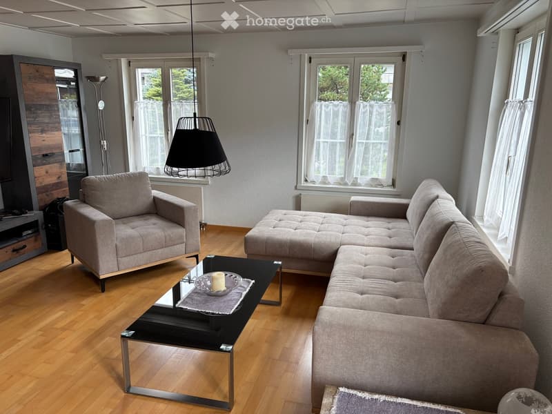 Modern living room with light grey sectional sofa, black coffee table, grey armchair, floor lamp, and TV stand. Large windows with white curtains and wooden flooring.