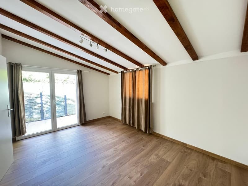 Empty attic room with wooden beams, wooden flooring, large sliding glass door with curtains, and recessed lighting.