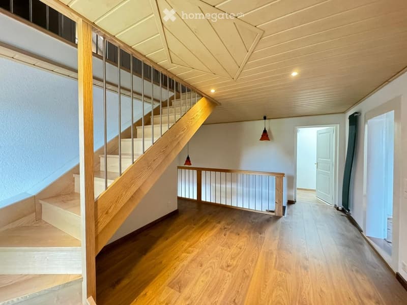 Spacious hallway with wooden flooring, a staircase with wooden steps and metal railing, and pendant lights hanging from the ceiling.