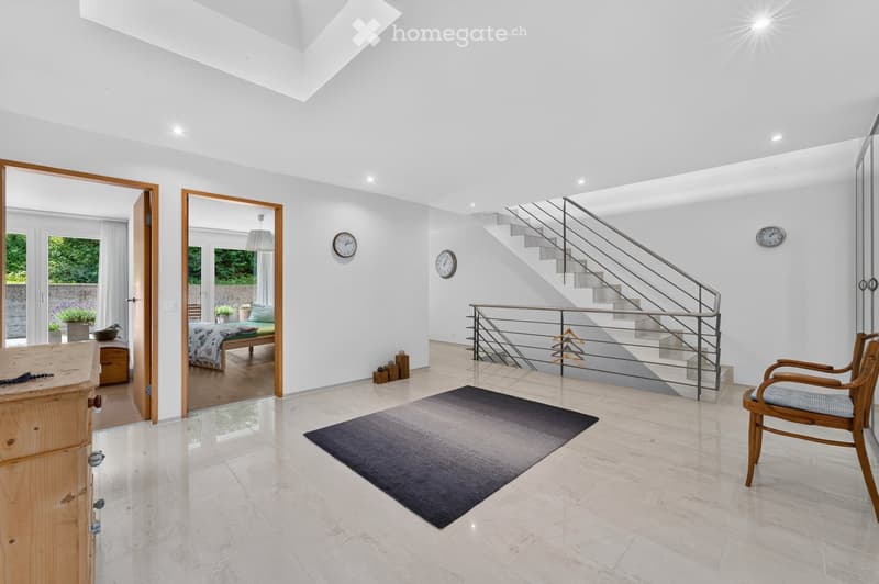 Bright, modern hallway with white walls, a staircase leading upwards, a wooden chair, a dark rug, and two wall-mounted clocks. The hallway features large windows and doors leading to other rooms.
