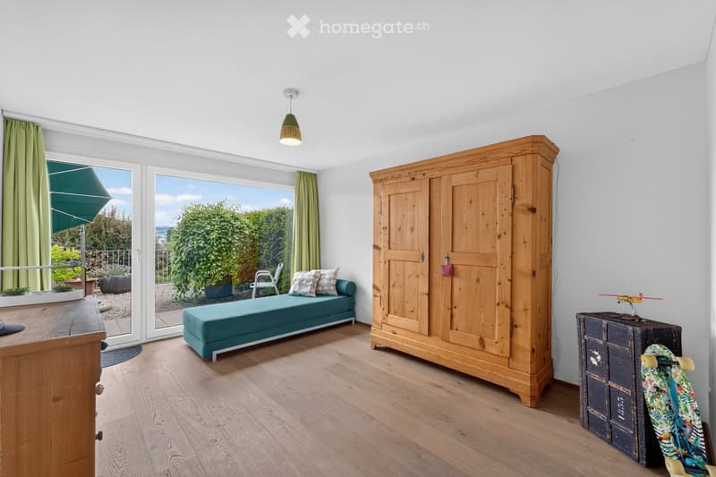 Bright room with wooden floor, large window offering a view, teal sofa, wooden wardrobe, skateboard, toy plane, and a small table.