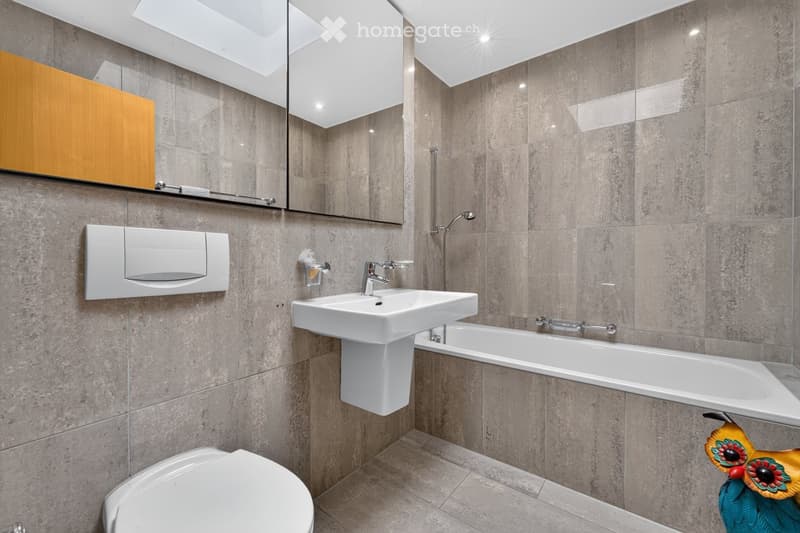 Modern bathroom with grey tiled walls and floor, white sink, bathtub, toilet, and a large mirror with wooden cabinet above it.