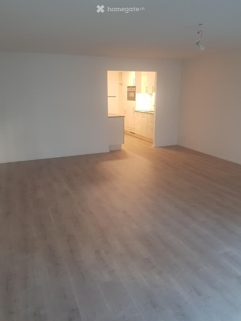 Empty room with wooden flooring and white walls, featuring an open doorway leading to a modern kitchen area with white cabinets and stainless steel appliances.