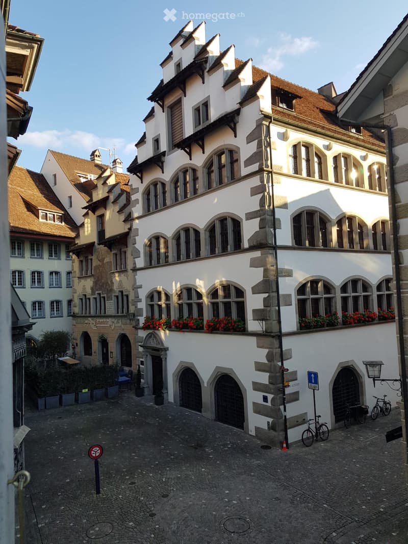 A historic white building with arched windows and a balcony adorned with red flowers, situated in a narrow cobblestone street lined with other traditional buildings.