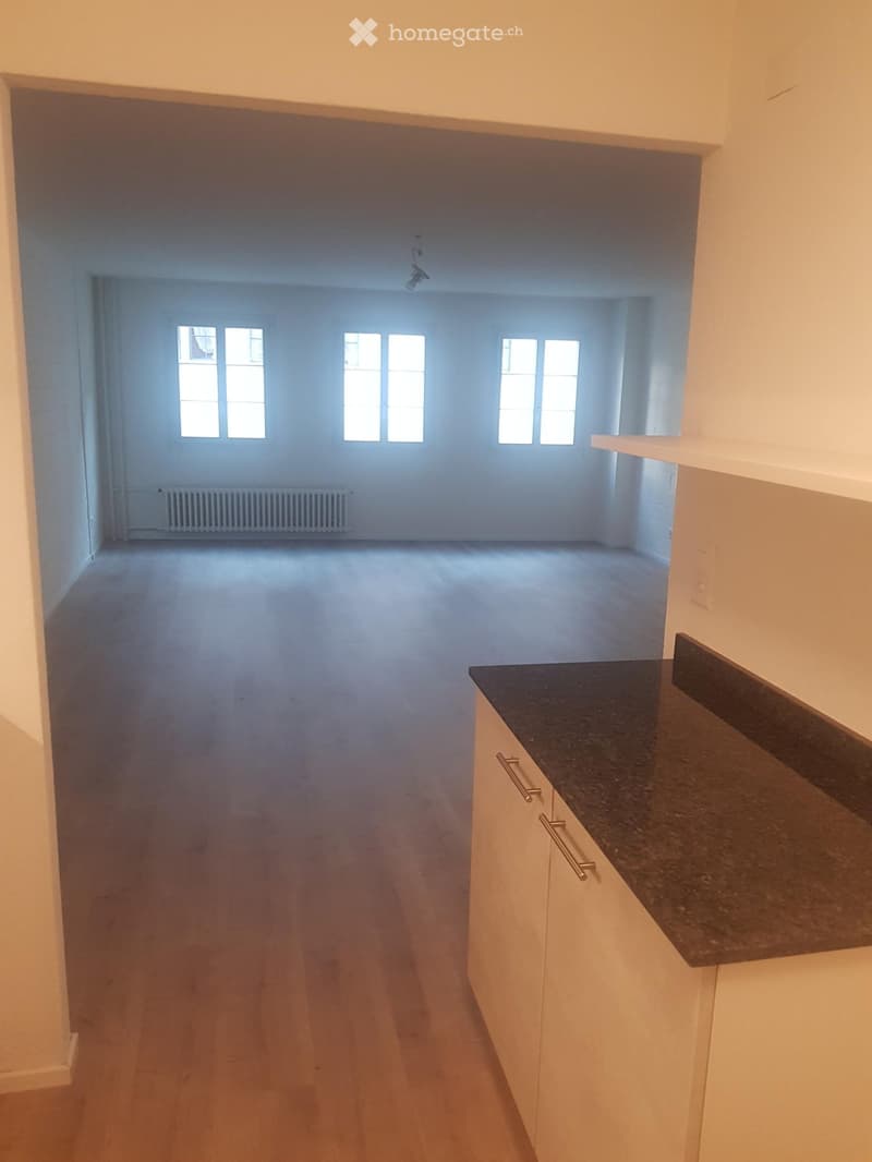 Empty room with wooden flooring, white walls, and large windows allowing natural light. A small kitchen area with white cabinets and a dark countertop is visible on the right side.