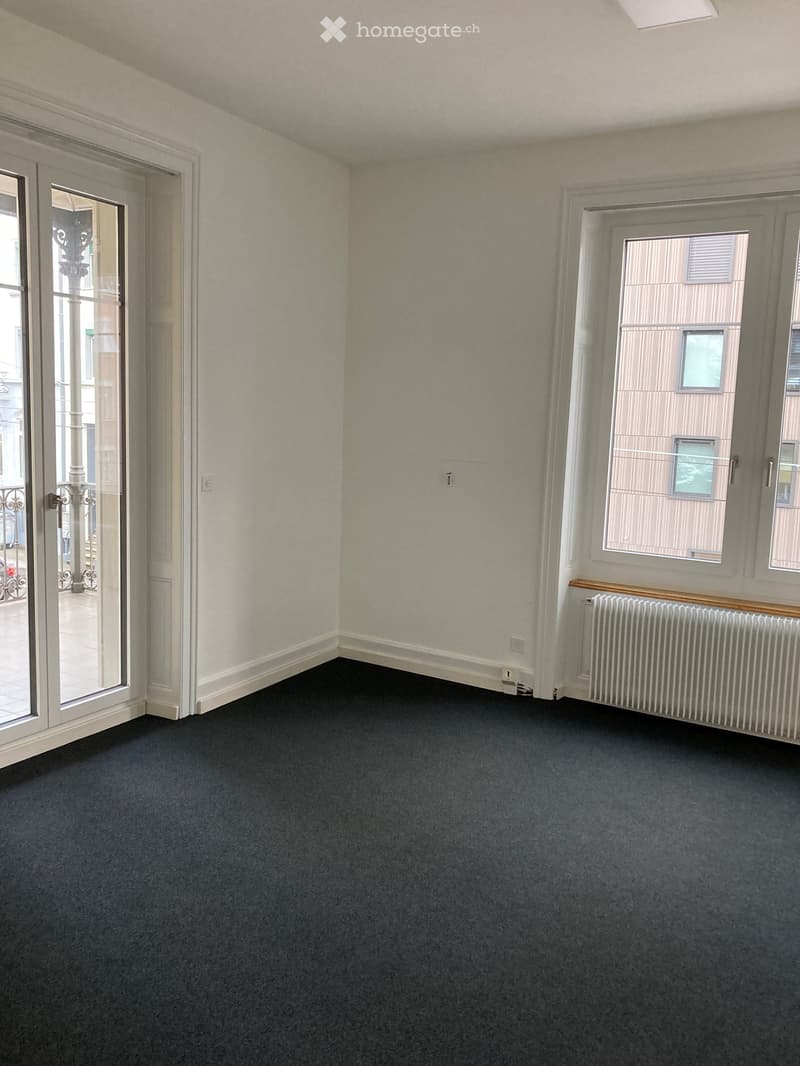 Empty room with dark carpet, white walls, large windows, radiator, and a sliding glass door leading to a balcony.