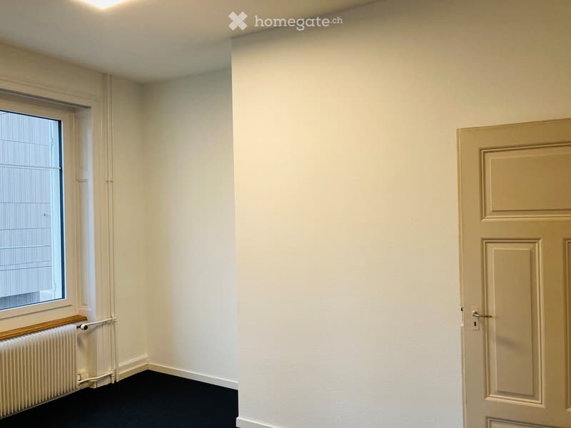 Empty room with white walls, black carpet, a window with blinds, a radiator, and a closed door.