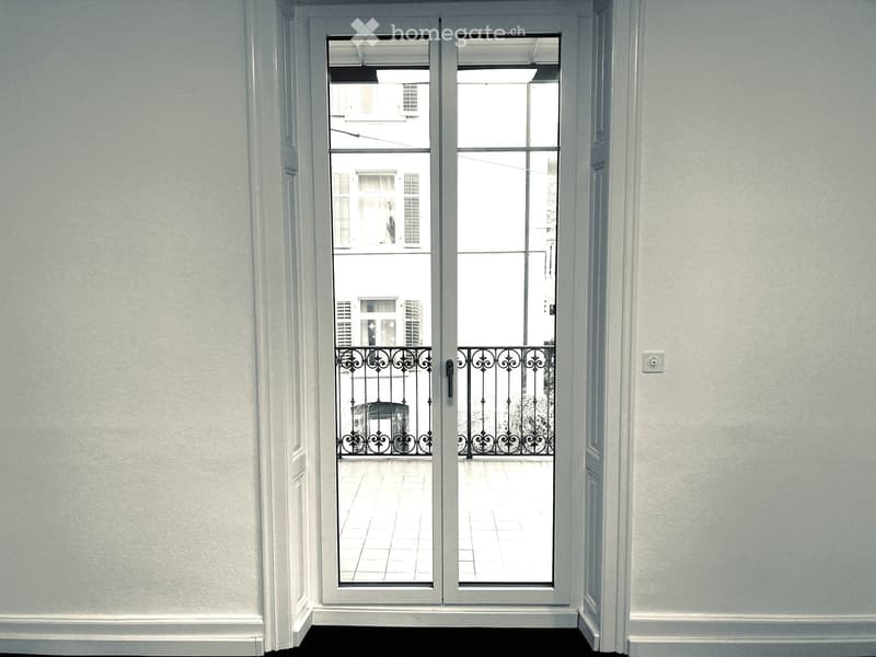 A hallway with a large glass door leading to a balcony with a decorative metal railing. The walls are light-colored, and the floor is dark.