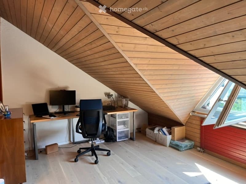 Bright attic room with wooden ceiling and floor, featuring a desk with a laptop, monitor, and chair, along with a lamp and storage unit.