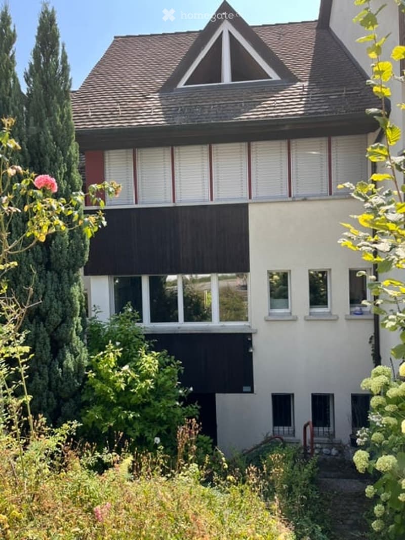 Two-story house with a triangular roof, white siding, multiple windows, and surrounding greenery.