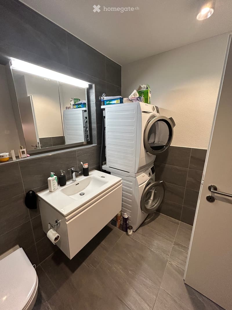 white toilet, white sink with faucet, stacked washing machine and dryer, mirror with lighting, white walls, wooden floor
