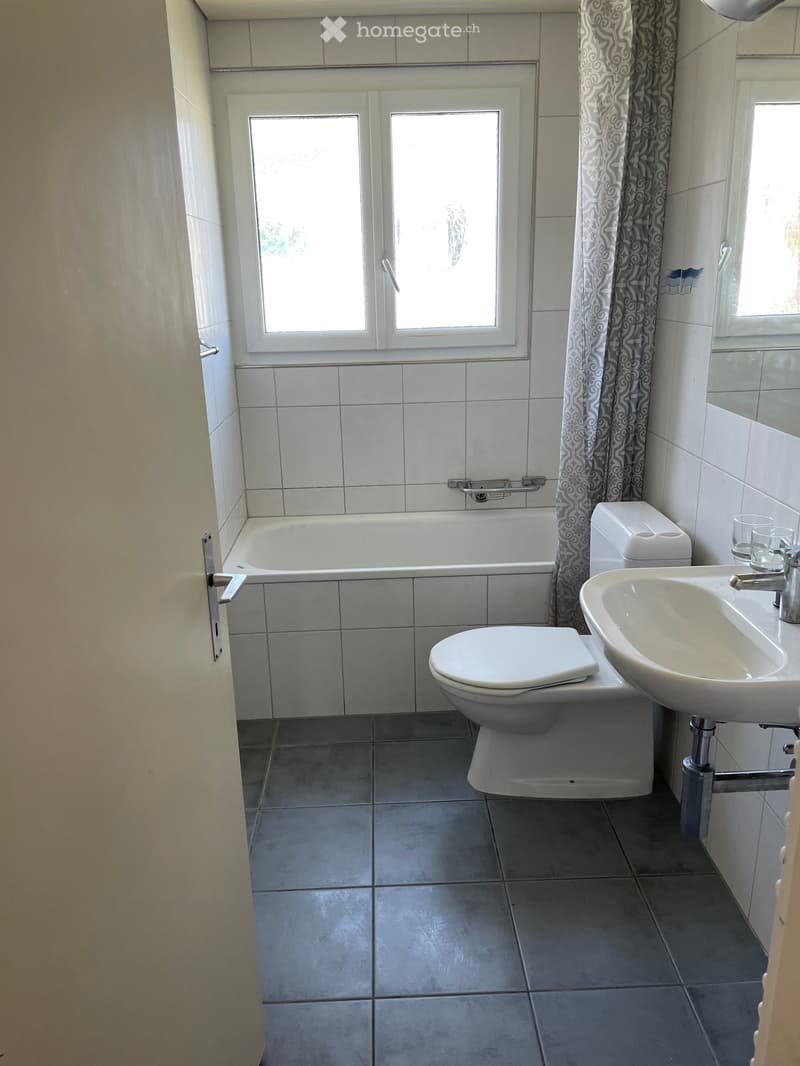 Bathroom with a bathtub, toilet, sink, and a window with a curtain. The room has tiled walls and floor.