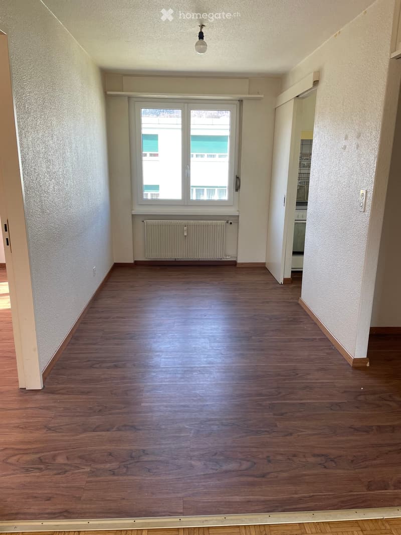 Empty hallway with wooden flooring, white walls, a window with a radiator below, and a doorway leading to another room.