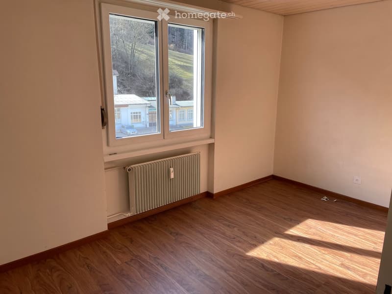 Empty room with wooden flooring, white walls, and large windows offering a view of the outdoors. A radiator is visible under the windows.