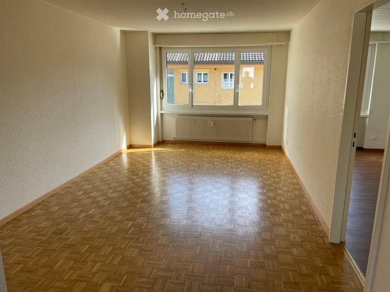 Empty room with wooden herringbone flooring, light-colored walls, and a large window with a radiator below it.