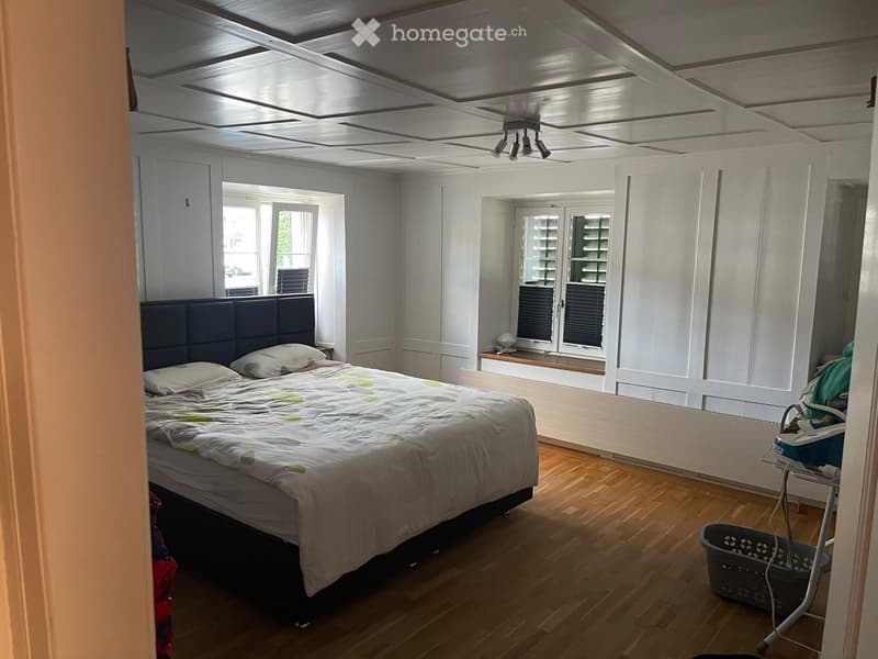 Bright bedroom with wooden flooring, large windows, a neatly made bed with a headboard, and a baby chair in the corner.