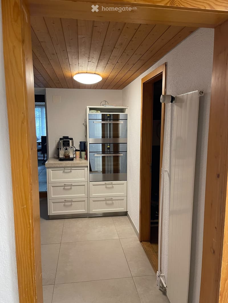 A narrow hallway with wooden ceiling beams, a recessed ceiling light, and a view into a modern kitchen area with a built-in oven, drawers, and a coffee machine on the counter.