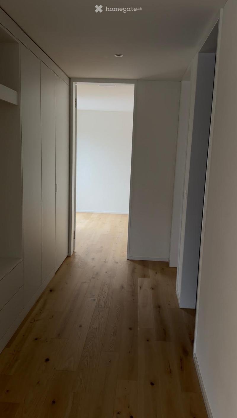 A modern, minimalist hallway with light wooden flooring and white walls, featuring built-in storage units on one side and an open doorway leading to another room.
