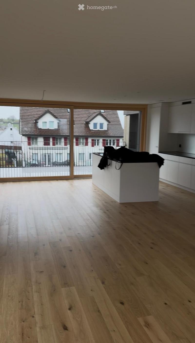 Empty room with light wooden flooring, white kitchen counter, and large windows overlooking a residential area.
