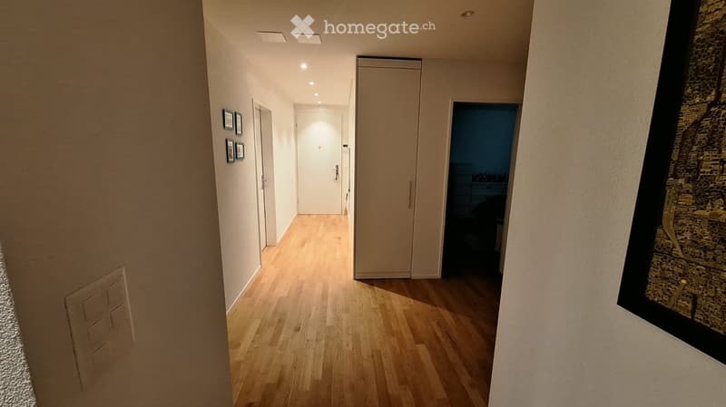 A narrow hallway with light wooden flooring, white walls, and recessed ceiling lighting. There are several doors and framed pictures on the walls.